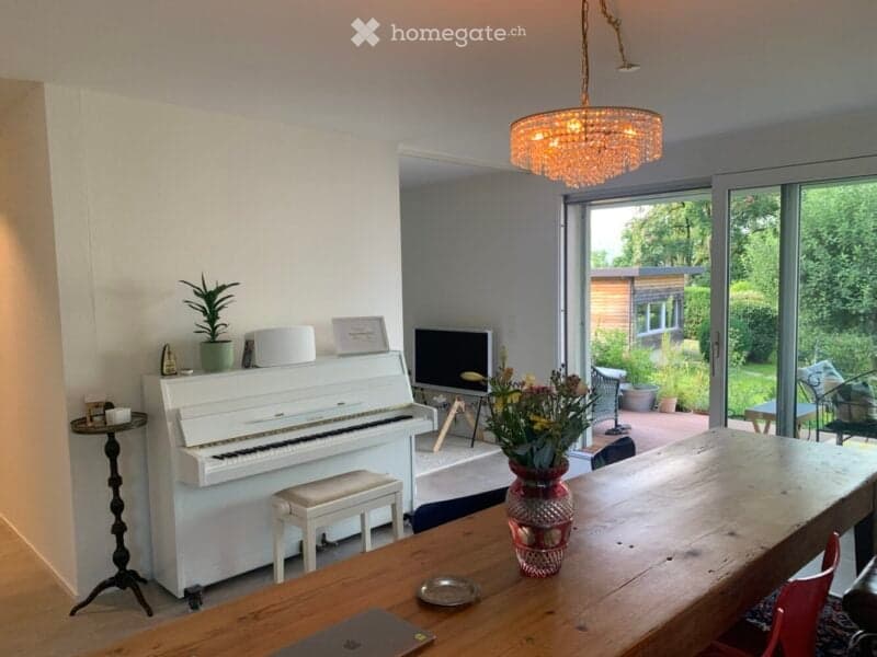 Bright living space with a white piano, wooden dining table, laptop, vase with flowers, and large windows offering a view of the garden.