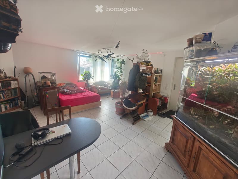 Well-lit room with a bed, sofa, fish tank, bookshelf, and a desk with a laptop and headphones. The room features tiled flooring and large windows with sheer curtains.