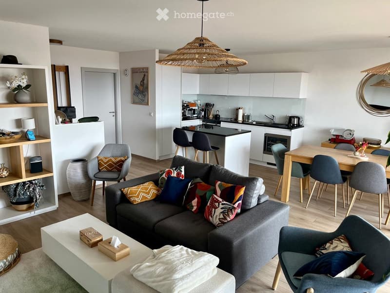 Modern open-plan living and dining area with a grey sofa, coffee table, dining table, and kitchen island. Features include a hanging wicker lamp, wooden flooring, and contemporary decor.