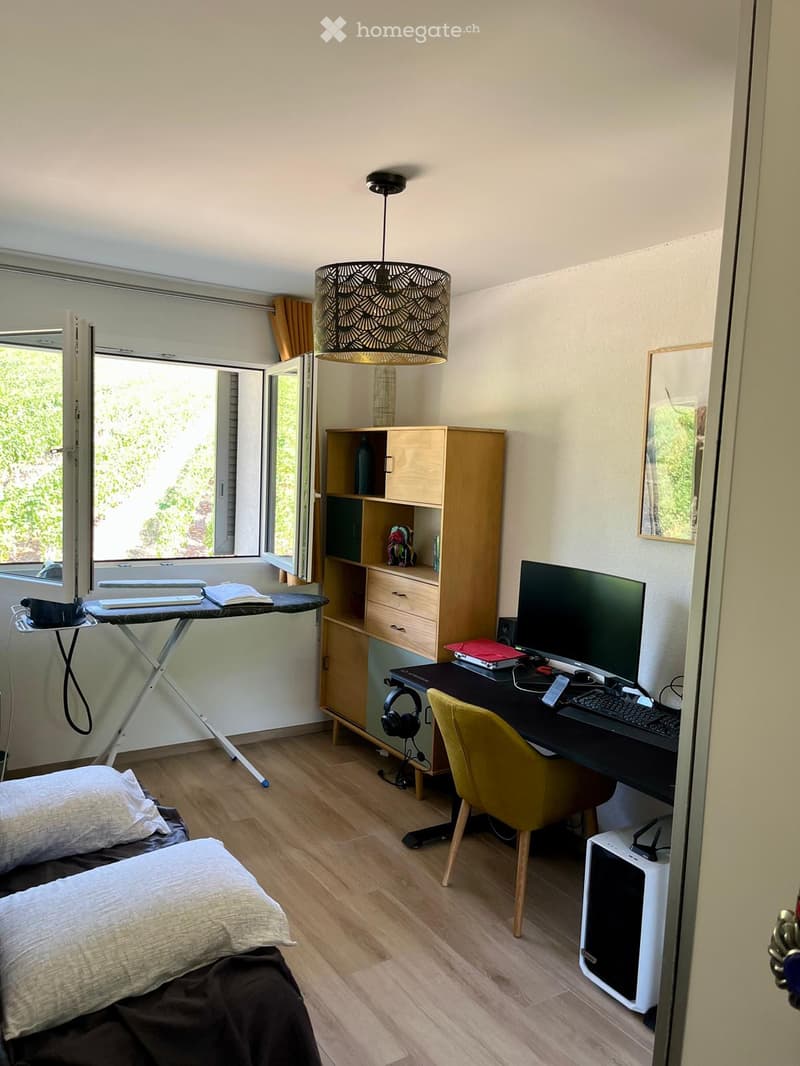 A multi-functional room with a bed, desk, and ironing board. Features a wooden floor, large window with blinds, and modern lighting.