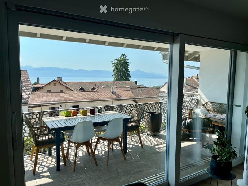 Balcony with a scenic view of a lake and mountains, featuring a wooden table, white chairs, a sofa, and potted plants.
