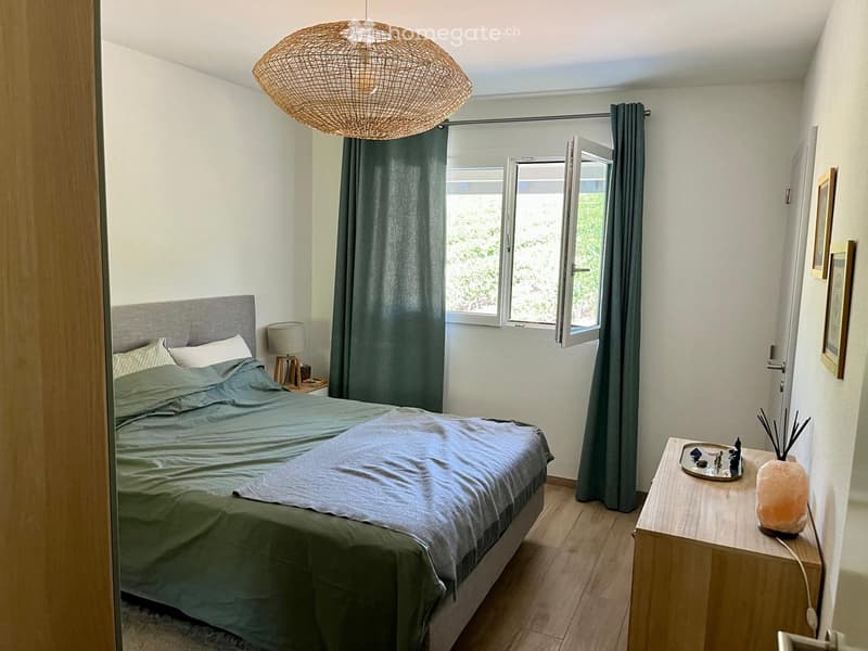 Cozy bedroom with a large bed, nightstand, lamp, and a window with open curtains. A decorative table with a salt lamp and small items is visible.