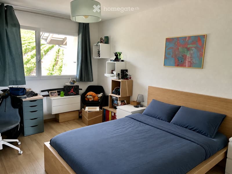 A modern bedroom with a wooden bed, blue bedding, a desk with a chair, shelves, and a large window with a view of greenery.