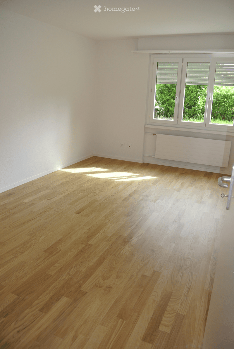 Empty room with light wooden flooring and white walls, featuring a large window with blinds and a radiator below it.