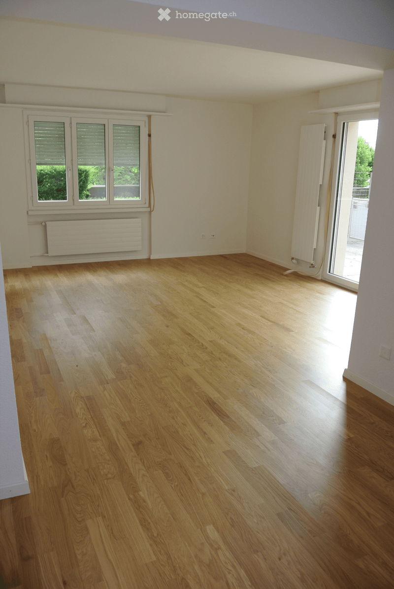 Empty room with wooden flooring, white walls, large windows, and a sliding glass door leading to a balcony.