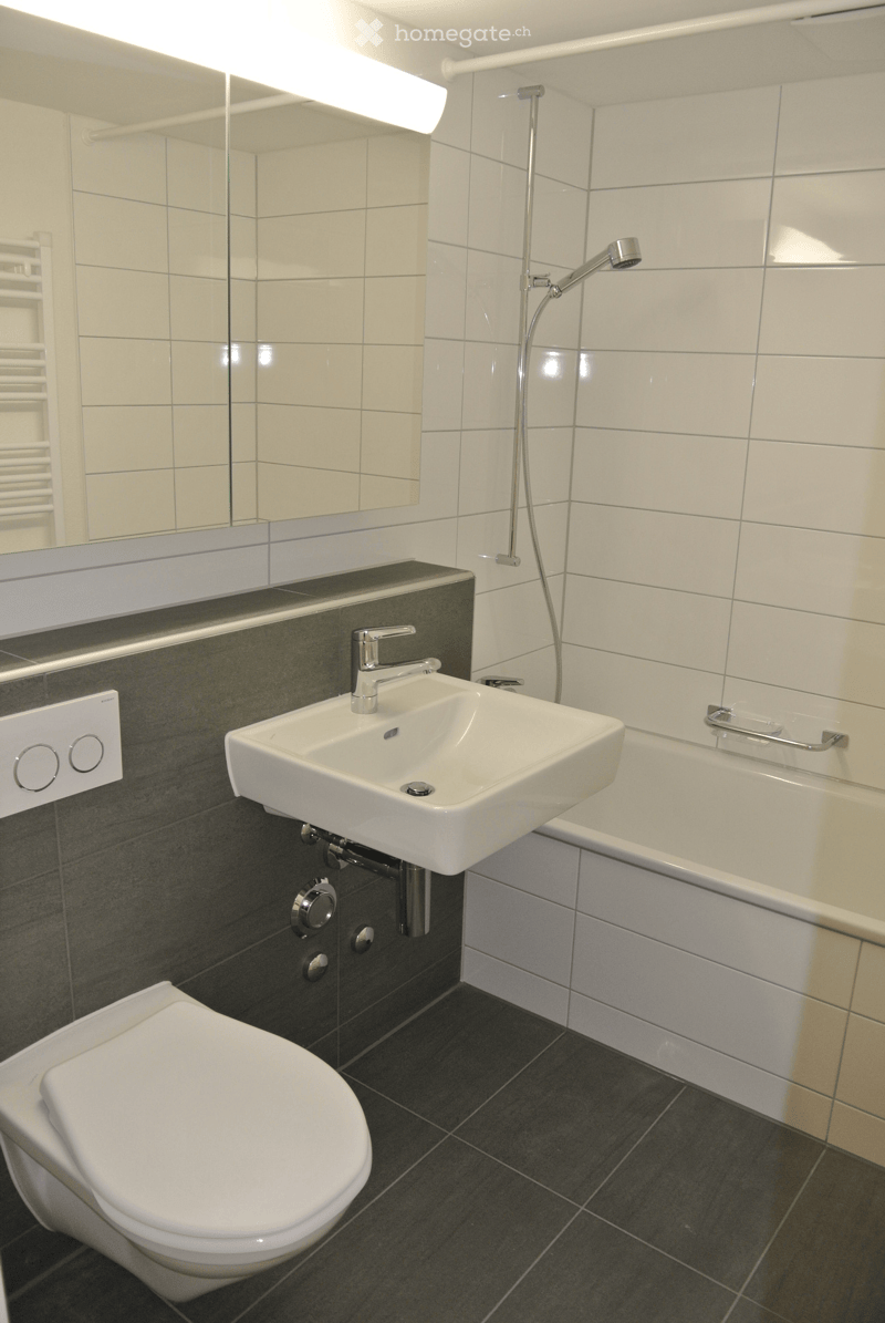 Modern bathroom with white tiles, dark grey accents, a toilet, a sink with a modern faucet, a bathtub, and a showerhead.
