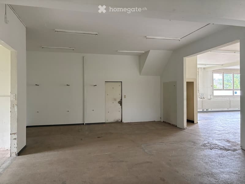 Empty room with bare walls, concrete floor, and a single door. Exposed ceiling beams and fluorescent lighting are visible.