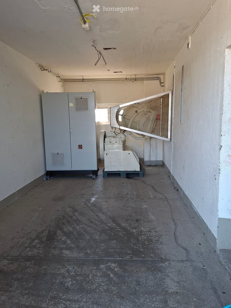 Empty room with concrete floor and white walls, featuring two large industrial machines, one with a fan and the other with a control panel.