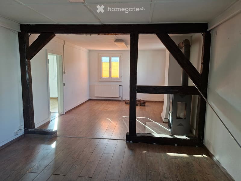 Empty room with wooden flooring, white walls, and a visible staircase leading to another level. There is a window with blinds, a radiator, and a small storage area with boxes.
