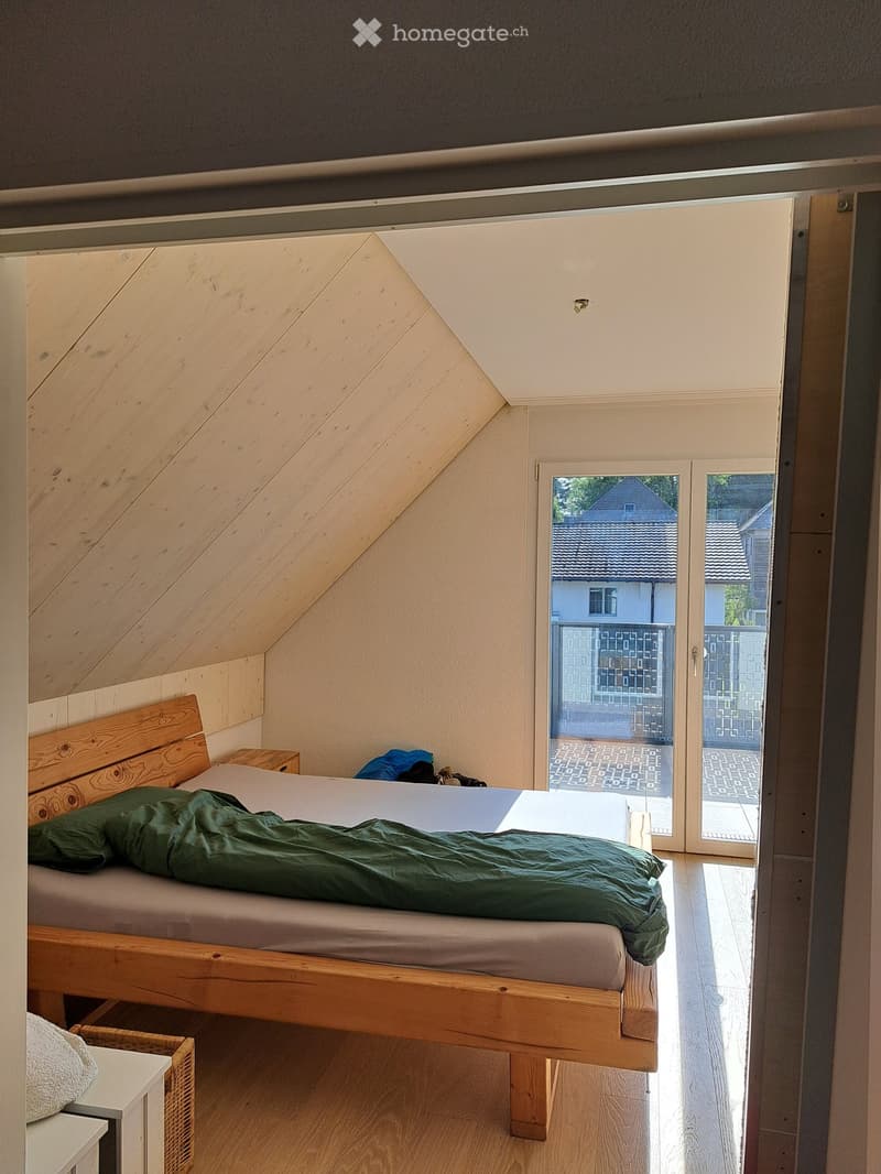 Cozy attic bedroom with wooden bed, nightstand, and wicker basket, featuring slanted wooden ceiling and large window with view of neighboring house.