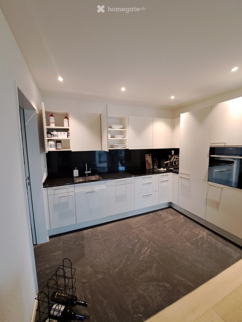 Modern kitchen with white cabinets, black countertops, stainless steel sink, and built-in oven. Includes a wine rack with several bottles, a coffee machine, and various kitchen utensils.