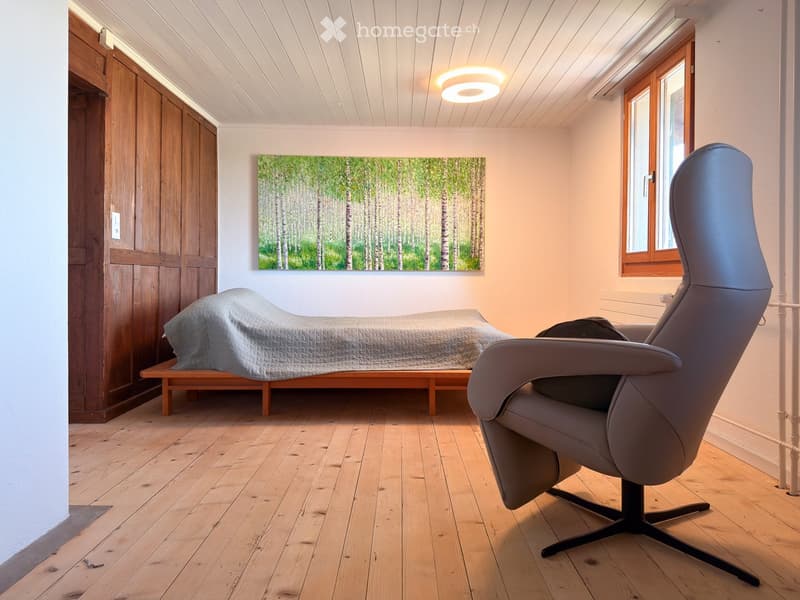 Cozy bedroom with wooden flooring, a bed with a grey duvet, a large armchair, and a painting of birch trees on the wall.