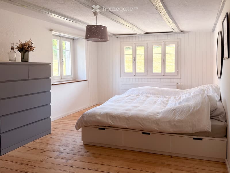 Bright bedroom with wooden floors, white walls, large windows, a bed with white bedding, a nightstand with a vase and bottle, and a hanging pendant light.