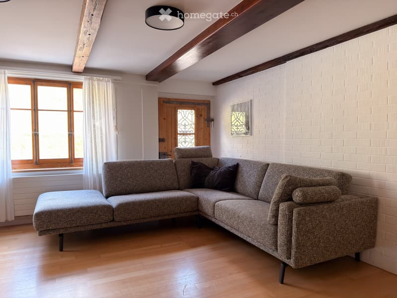 Cozy living room with exposed wooden beams, a grey sectional sofa with dark cushions, wooden flooring, and large windows with white curtains.