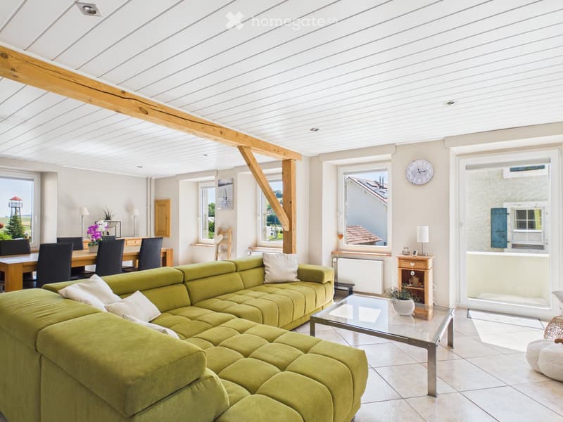 Bright living room with a green sectional sofa, glass coffee table, dining area with chairs and table, large windows, and decorative items.
