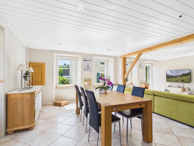 Bright, open-plan living and dining area with a wooden dining table, chairs, a green sofa, a lamp, and a large TV screen displaying a scenic image.