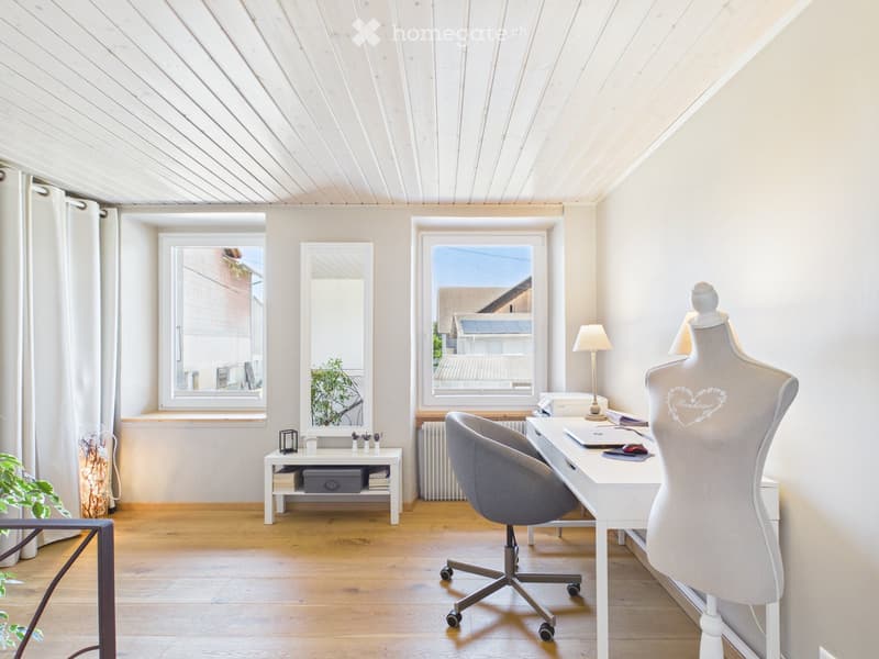 Bright, modern room with wooden flooring, white walls, and slanted ceiling. Features a desk with a laptop, a chair, a mannequin, a small table with books and a lamp, and a large window with a view of the roof.