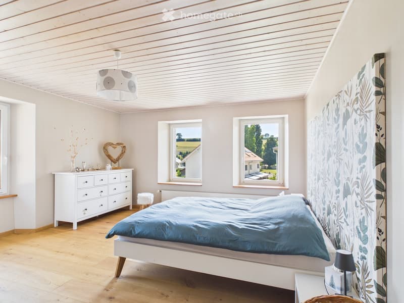 Bright bedroom with wooden flooring, white walls, and large windows offering a view of the landscape. Features a blue duvet-covered bed, white dresser, nightstand with lamp, and decorative items.