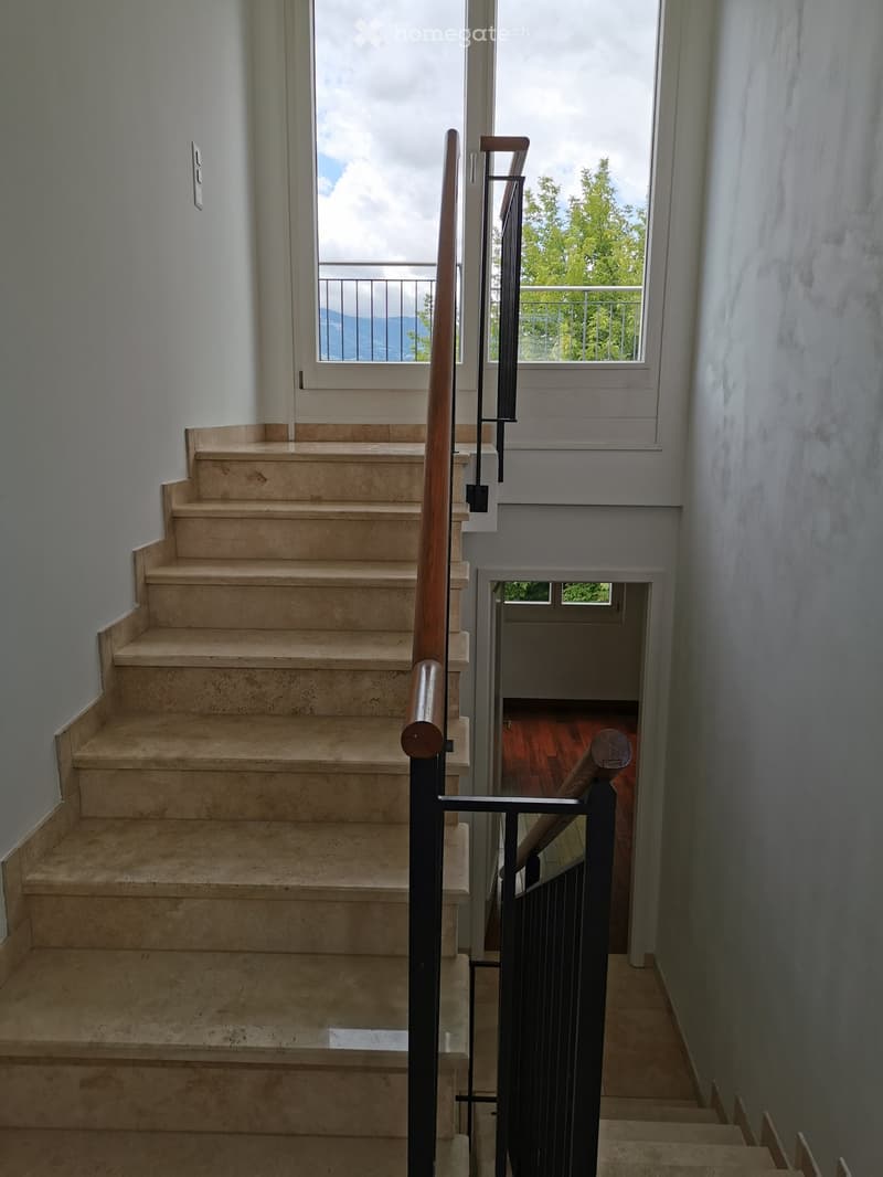 Staircase with beige stone steps, wooden handrail, and metal balustrade, leading to a room with wooden flooring visible at the end.