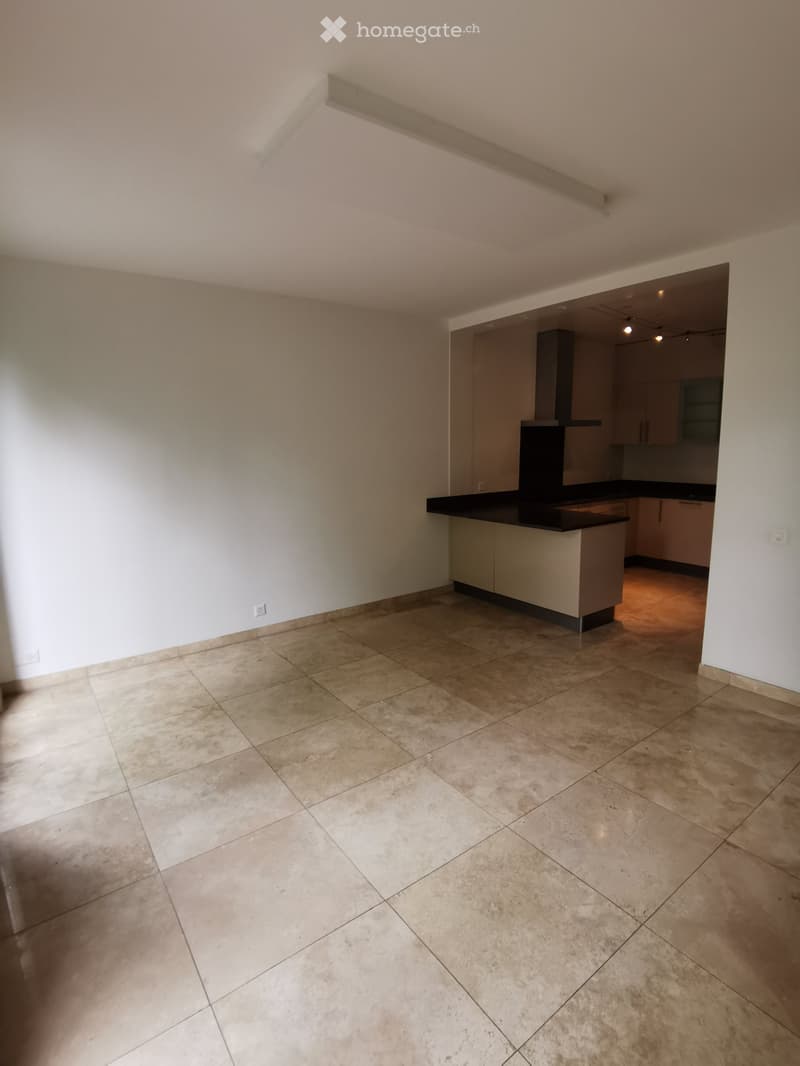 Empty room with tiled floor and white walls, featuring a small kitchen area with cabinets and a countertop in the background.