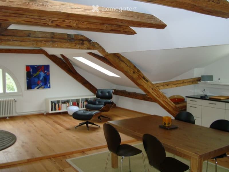 Open-plan living and dining area with exposed wooden beams, hardwood flooring, a black leather sofa, a dining table with chairs, a bookshelf, and a small kitchen area with cabinets.