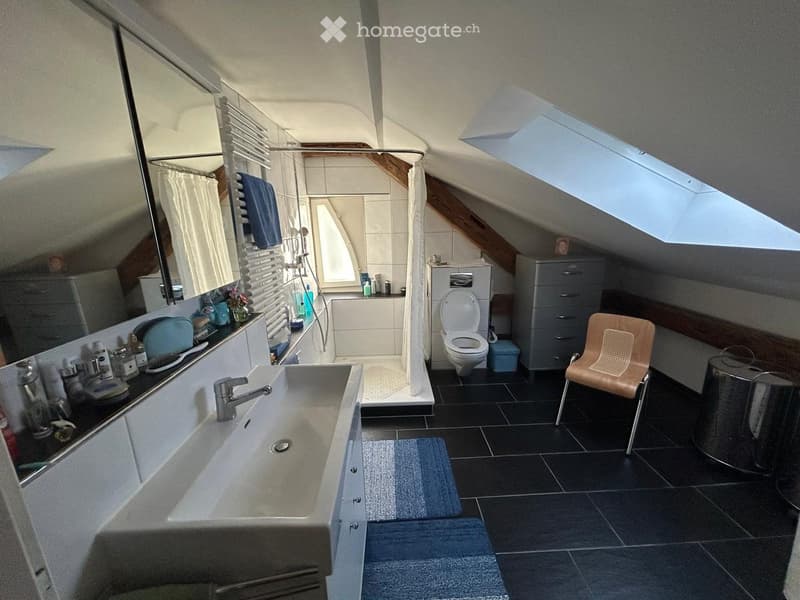 Modern attic bathroom with skylight, dark tiled floor, white sink, toilet, bathtub, and a small wooden chair.