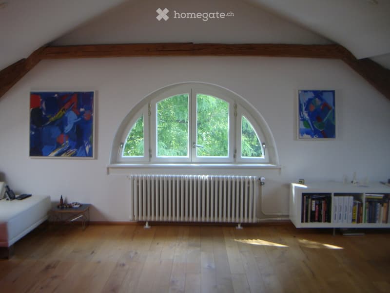 Bright attic room with wooden floors, large arched window, white radiator, bookshelf, small table, and paintings on the walls.