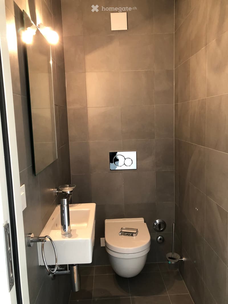 Modern bathroom with a white sink, chrome faucet, toilet, and a mirror with built-in lighting. The walls are tiled in a neutral color, and the floor is dark.