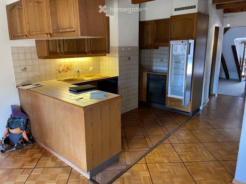 Well-lit kitchen with wooden cabinets, a central island, and modern appliances including a refrigerator and oven. The island features a tiled countertop with a sink and various items, including a backpack and papers.