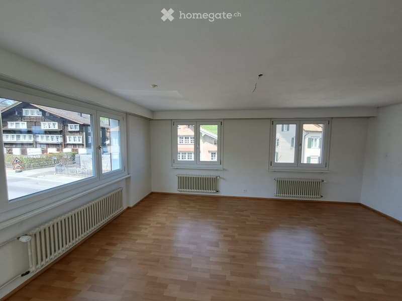 Empty room with wooden flooring, white walls, and multiple windows offering a view of the street and neighboring houses. Radiators are visible along the walls.