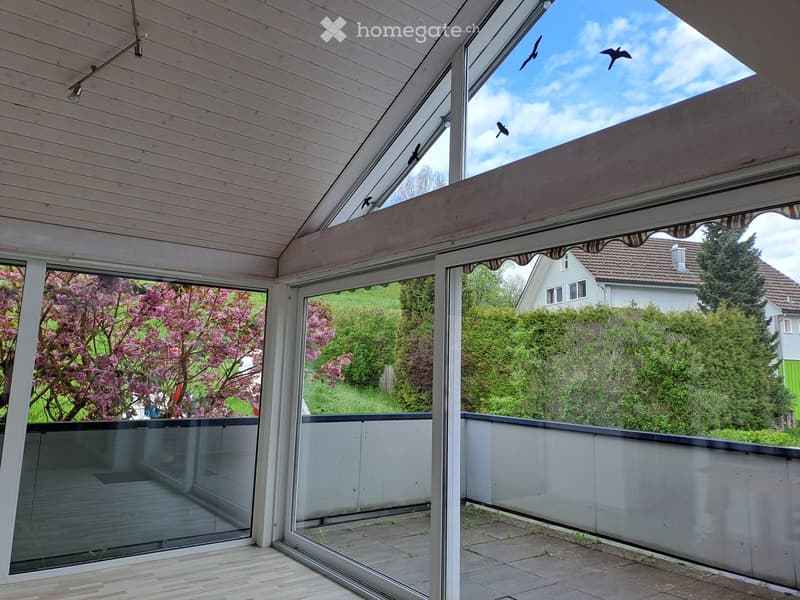 Spacious balcony with wooden ceiling, glass railings, and open views of a garden and neighboring house.