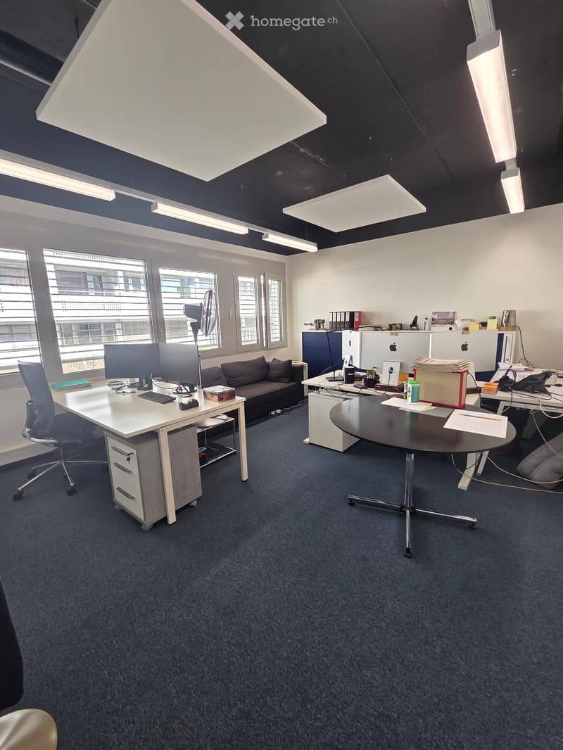 Modern office space with desks, chairs, a sofa, and various office equipment. Features large windows with blinds and a fan near the window.