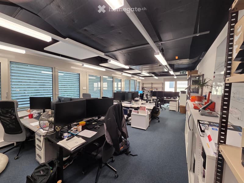 Office space with desks, chairs, multiple computer monitors, keyboards, mice, fans, cups, backpacks, and a plant. The room has large windows with blinds and exposed ceiling beams.