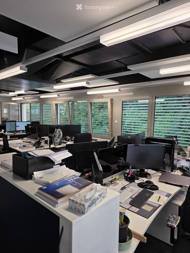 Modern office space with multiple desks, computers, chairs, and office supplies. Large windows provide natural light, and the room is equipped with various office equipment and accessories.