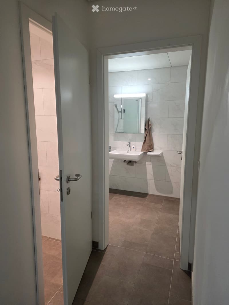 Modern bathroom with a white sink, large mirror, and a towel hanging on the side. The floor is tiled in a warm brown color, and the walls are covered with light-colored tiles.