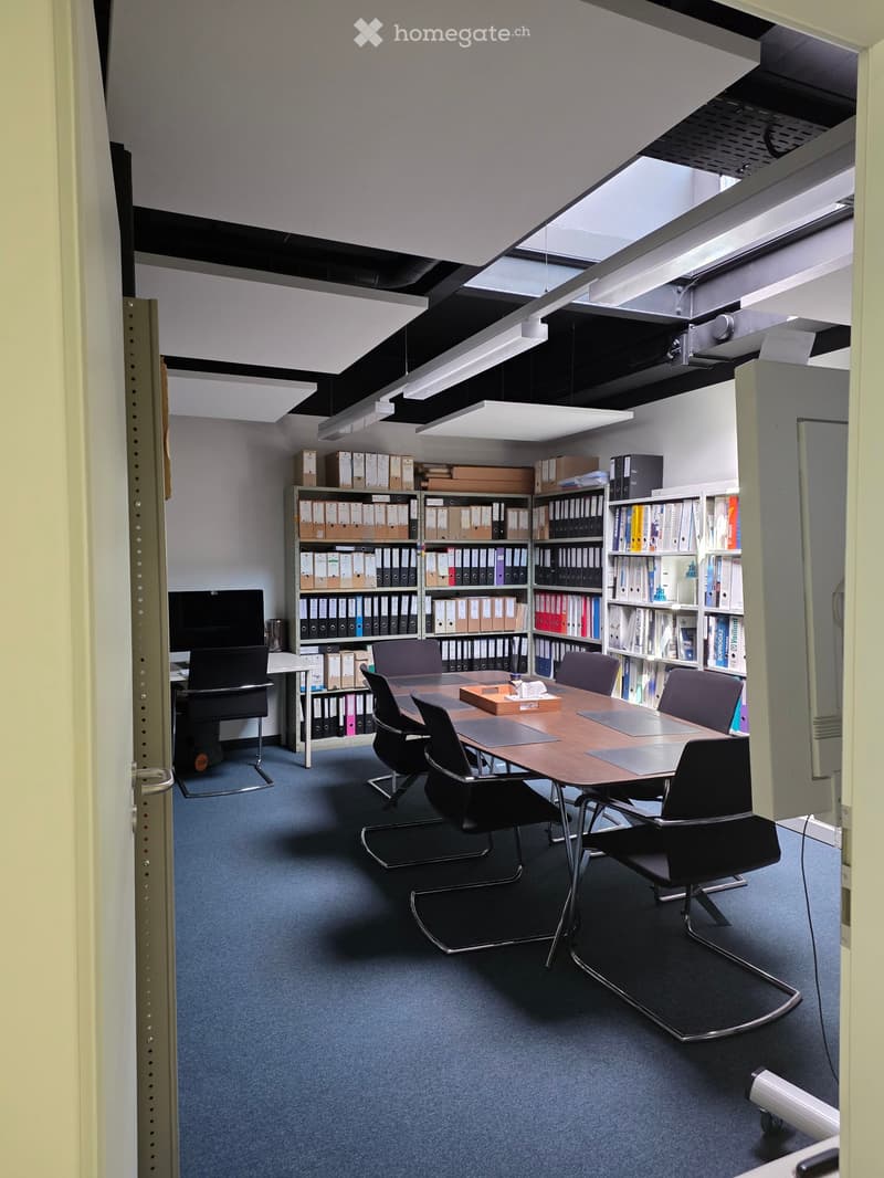 Office space with a large conference table, chairs, shelves filled with binders and books, and a computer screen.