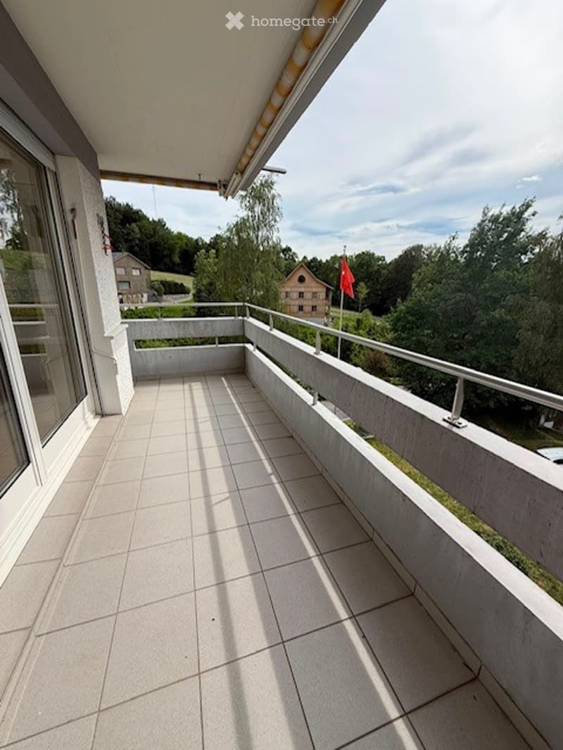 Spacious balcony with tiled flooring, white railings, and a scenic view of a house and greenery in the background.