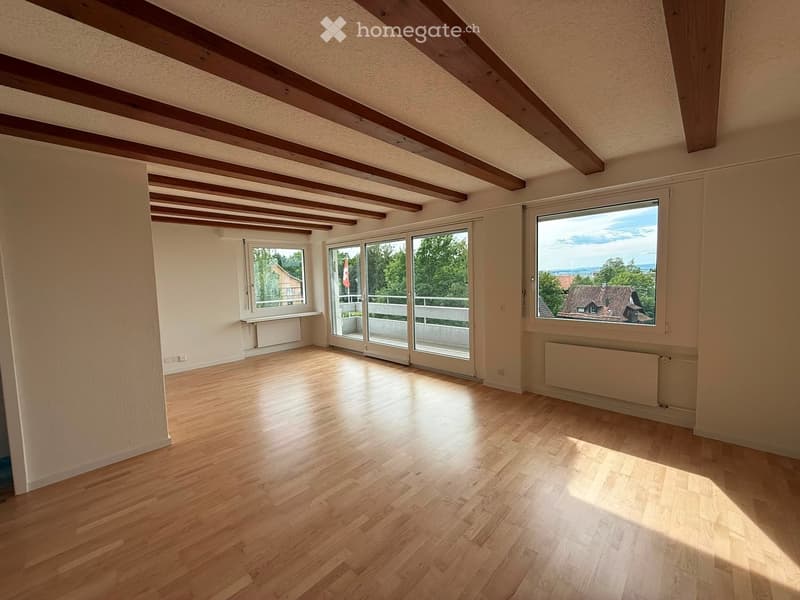 Empty room with wooden flooring, exposed wooden beams on the ceiling, large windows, and sliding glass doors leading to a balcony.