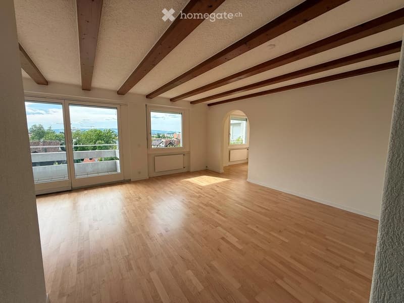 Empty room with wooden flooring, exposed wooden beams on the ceiling, large windows, and a sliding glass door leading to a balcony.