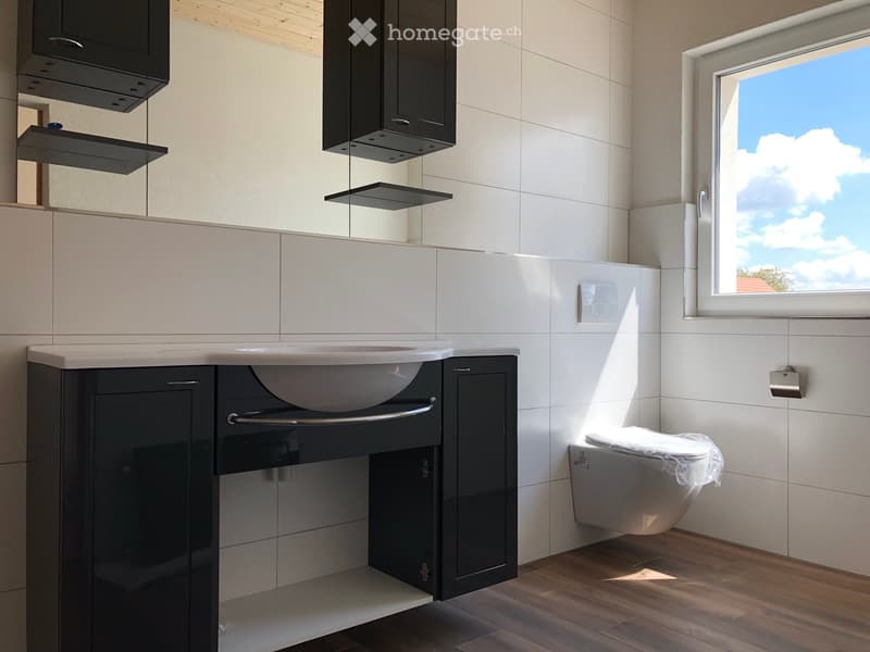 Modern bathroom with white tiles, dark cabinets, a sink, and a toilet. Features a large window with a view of the sky.