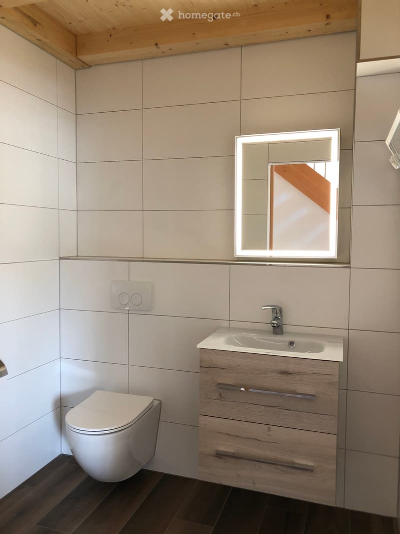 Modern bathroom with white tiled walls, a wooden vanity with a sink, a toilet, and a large illuminated mirror above the sink.