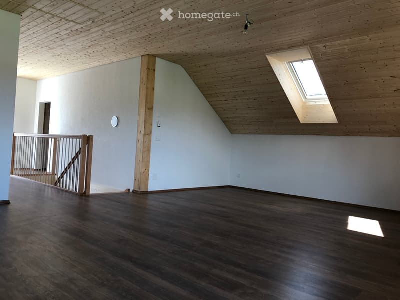 Empty room with wooden floor, white walls, wooden ceiling, and a skylight window. A staircase with a wooden railing is visible on the left side.