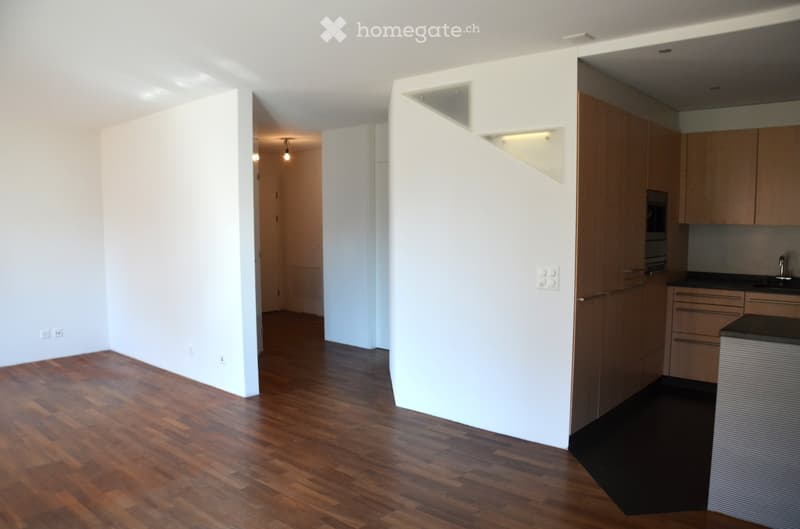 Empty room with wooden flooring, white walls, and a small kitchen area with cabinets and a sink.