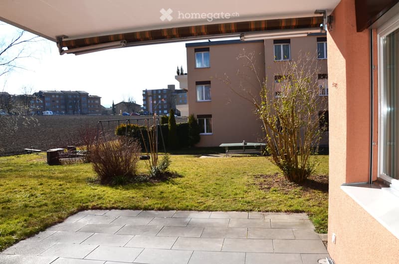 Well-maintained garden area with a paved pathway, grassy lawn, small plants, and a bench. The area is enclosed by a building with a covered walkway and overlooks a residential neighborhood.