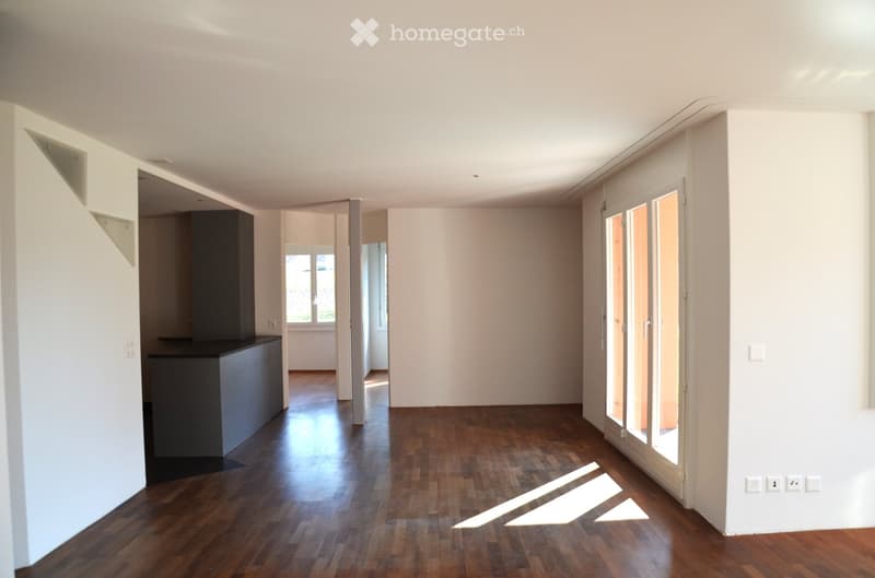 Empty room with wooden flooring, white walls, and large windows allowing natural light to enter. The room features a modern design with a sleek black cabinet visible in the background.