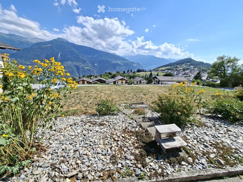 Garden area with gravel ground, small wooden bench, yellow flowers, and distant mountain view with houses and trees.