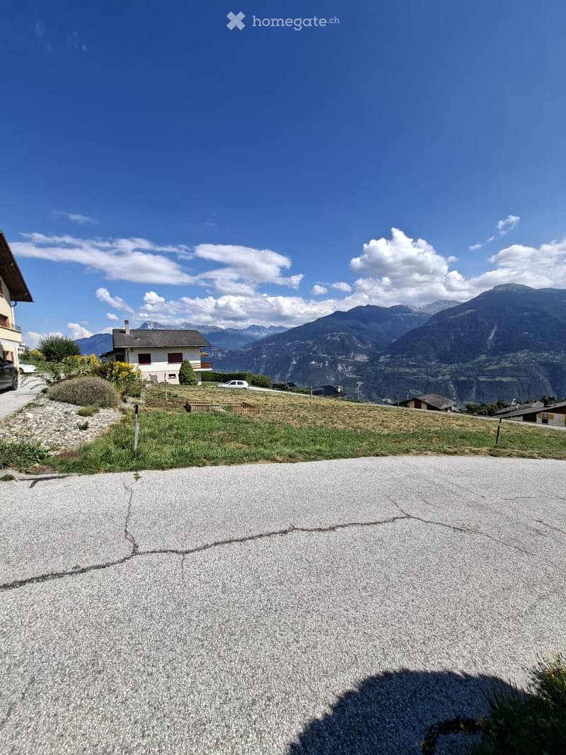 A gravel driveway leads up to a house with a view of mountains in the background. The house is surrounded by a small lawn and some shrubs.