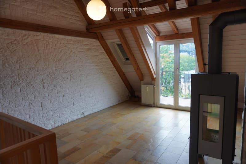 Empty attic room with exposed wooden beams, stone wall, tiled floor, large skylight, and a fireplace with a chimney. The room has a door leading to a balcony with outdoor greenery visible.