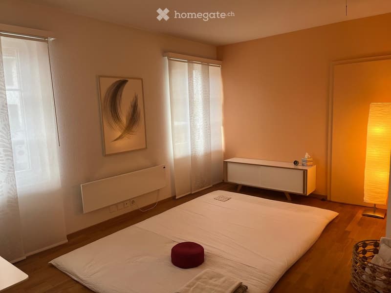 Minimalist bedroom with a large bed, a nightstand with a lamp, a picture frame on the wall, and a small stool on the bed.