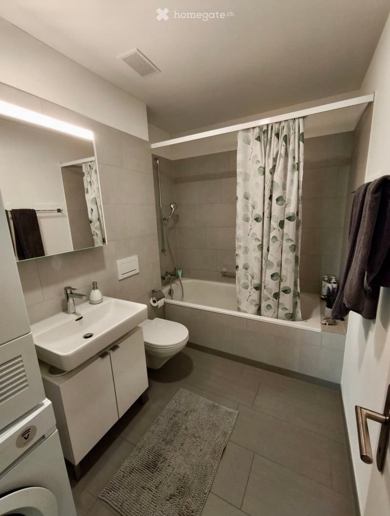 Modern bathroom with a white sink, toilet, bathtub, and shower area with a leafy-patterned curtain. Includes a mirror, towel, and a small rug on the floor.
